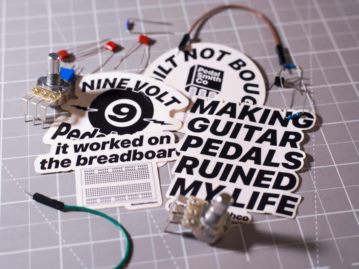 A DIY guitar pedal circuit board kit with various electronic components and printed stickers on a gray gridded cutting mat. The kit includes a potentiometer, transistors, resistors, a breadboard, and humorous stickers reading 'NINE VOLT', 'Making Guitar Pedals Ruined My Life', and 'It worked on the breadboard'.