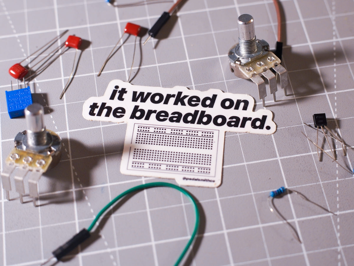 A sticker with the text 'it worked on the breadboard.' placed over a breadboard layout on a gray gridded cutting mat, surrounded by electronic components like potentiometers, LEDs, and transistors.