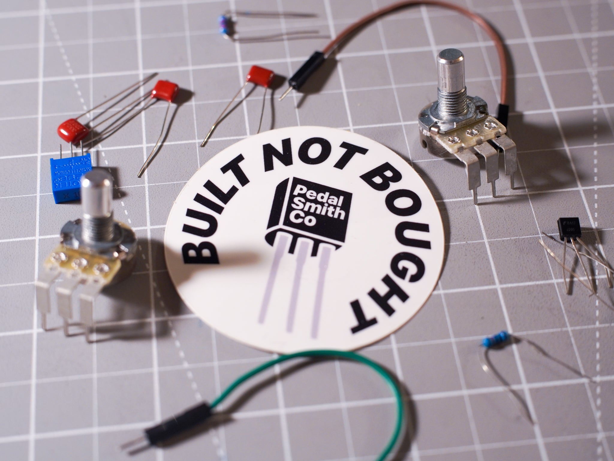 A collection of electronic components, including potentiometers, diodes, and transistors, arranged around a circular white sticker that reads 'BUILT NOT BOUGHT' with the Pedal Smith Co logo, placed on a gray gridded cutting mat.