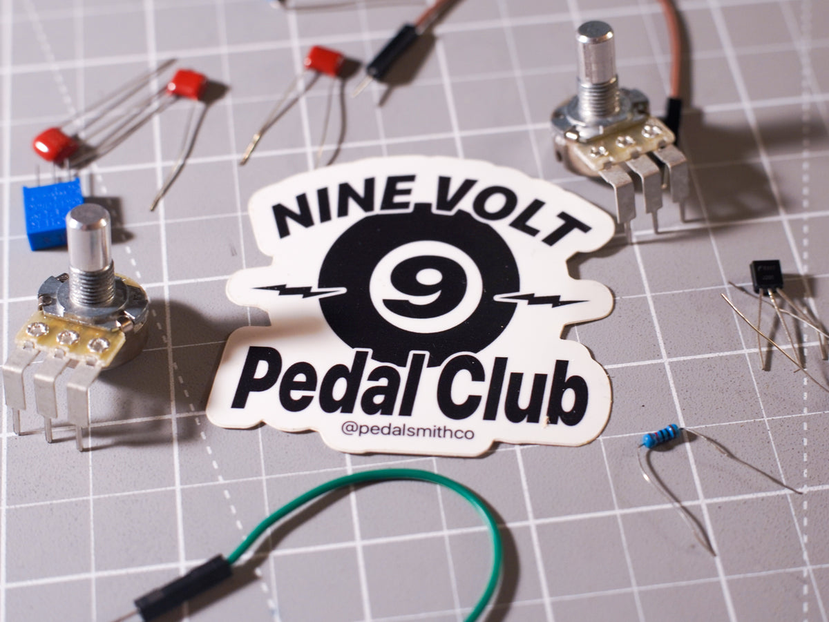 A die-cut sticker reading 'NINE VOLT Pedal Club' with a large black '9' in the center, placed on a gray gridded work mat surrounded by electronic components including potentiometers, capacitors, and a blue resistor.
