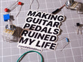 A die-cut sticker with the text 'MAKING GUITAR PEDALS RUINED MY LIFE' in bold black letters on a white background, placed on a gray gridded work surface surrounded by electronic components.