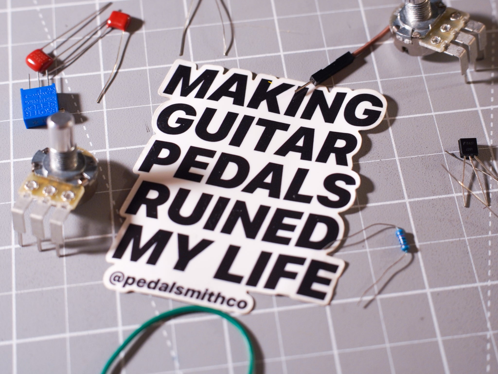 A die-cut sticker with the text 'MAKING GUITAR PEDALS RUINED MY LIFE' in bold black letters on a white background, placed on a gray gridded work surface surrounded by electronic components.